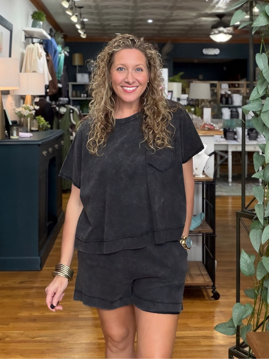 A woman with curly blonde hair stands in a boutique wearing a matching black washed short-sleeve t-shirt and shorts set. The background shows a retail store interior with wooden floors, shelves, and plants.