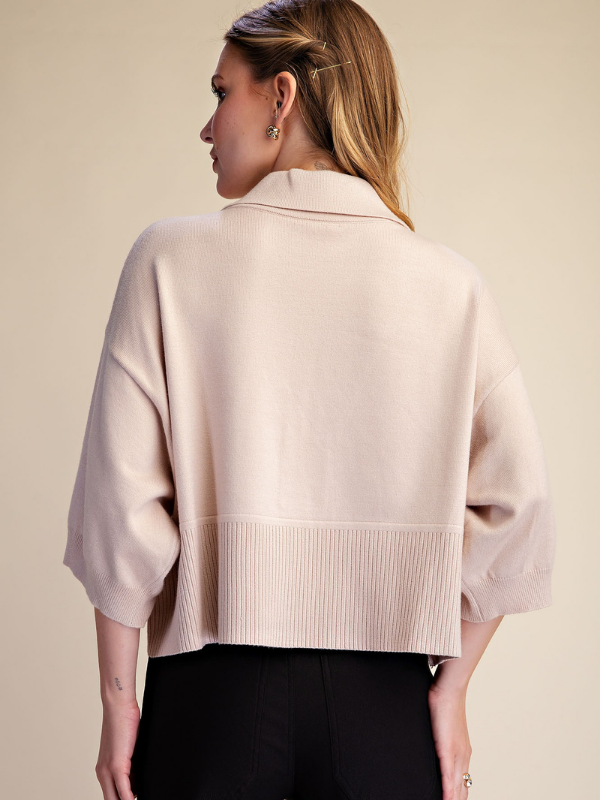 A woman shown from the back wearing a light beige knit cropped sweater with three-quarter sleeves and a ribbed hem, paired with black pants, against a solid beige background.