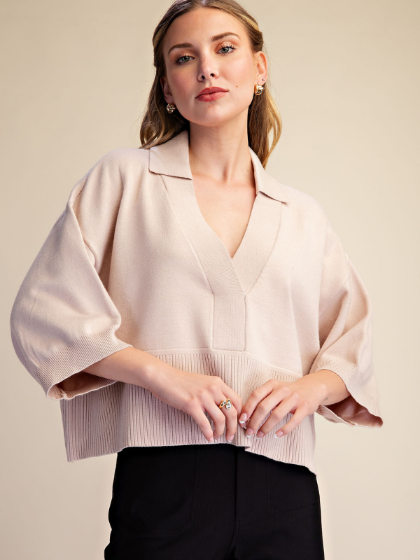 A woman wearing a light beige cropped blouse with wide balloon sleeves and a V-neckline, paired with black trousers, against a solid beige background.