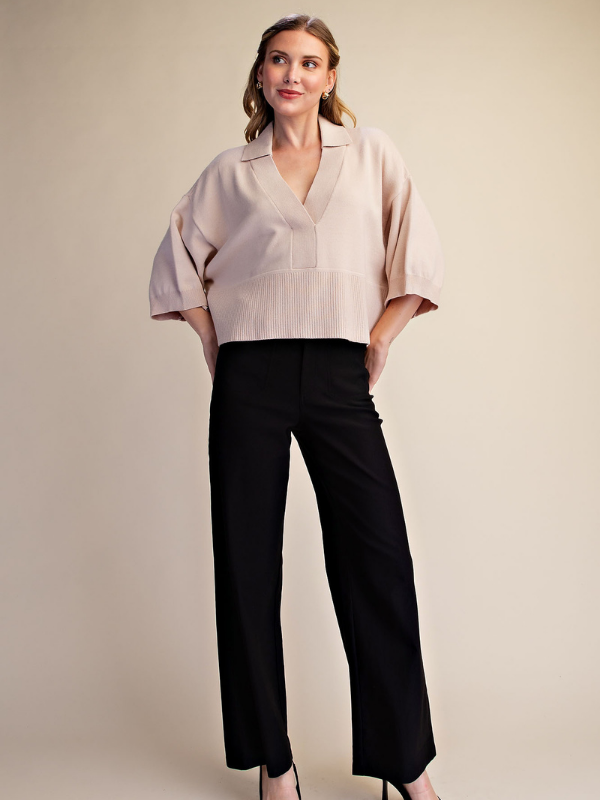 A woman modeling a light beige short-sleeve blouse with a V-neck and black wide-leg trousers, standing against a plain beige studio background.