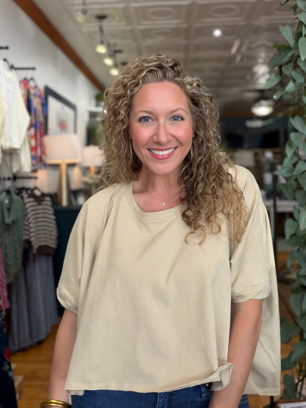 A smiling woman with curly blonde hair wearing a loose, short-sleeve beige top and blue jeans, standing inside a boutique store.