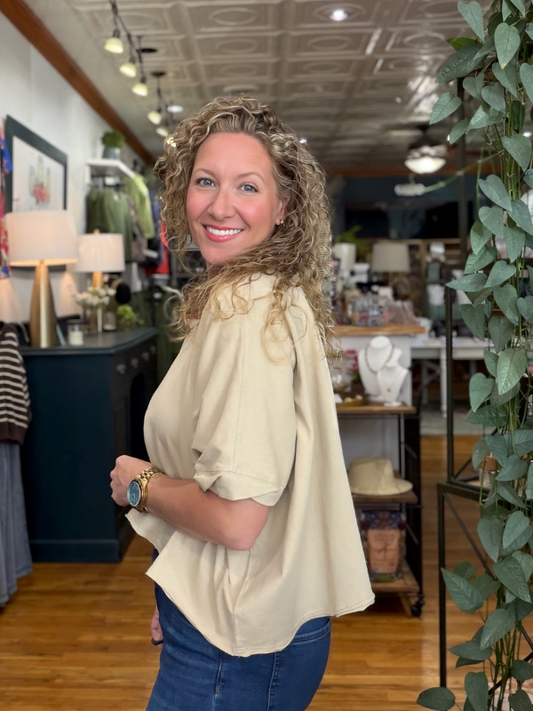 A smiling woman with curly blonde hair wearing a beige short-sleeve blouse and blue jeans, standing inside a retail store with wood floors, shelves, and plants in the background.