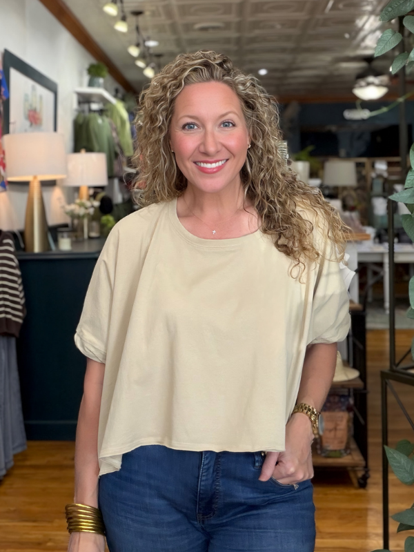 A smiling woman with curly blonde hair wearing a light beige short-sleeve crop top and blue jeans, standing inside a retail store.