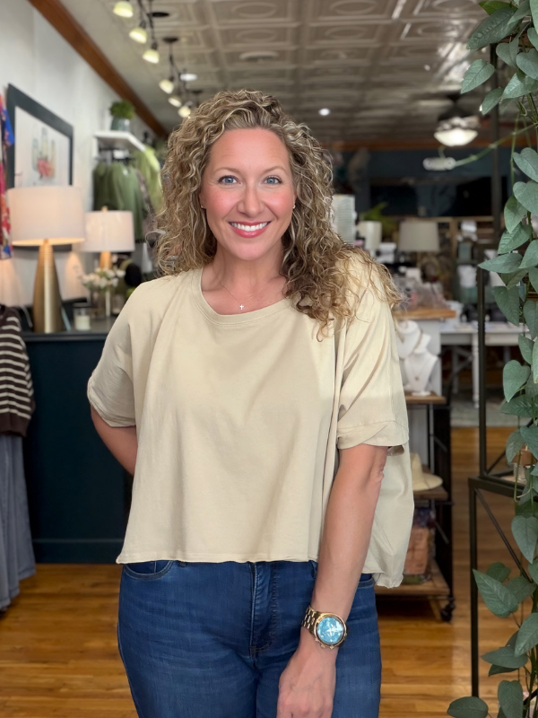 A smiling woman with curly blonde hair stands in a boutique wearing a light beige short-sleeve crop top and blue jeans.