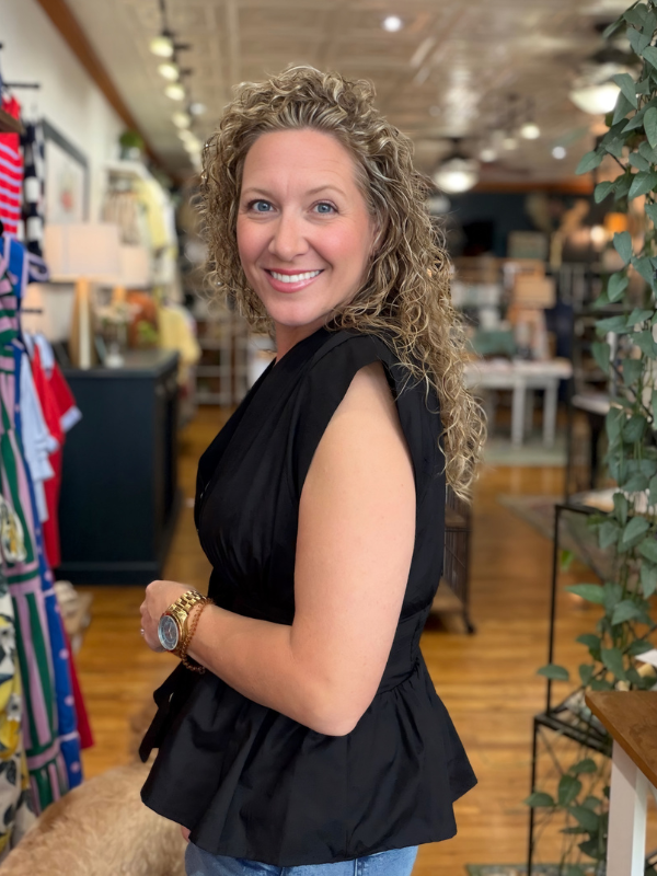 Woman in a black sleeveless top standing in a store with clothing and decor in the background.