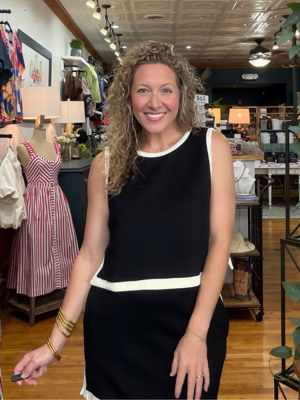 A smiling woman with curly hair stands in a boutique wearing a sleeveless black dress with white contrast trim at the neckline, waist, and hem. The background shows clothing racks, mannequins, lamps, and store fixtures.
