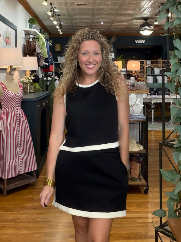 A smiling woman with curly blonde hair stands in a boutique wearing a sleeveless black mini dress with contrasting white trim at the neckline, waist, and hem. The background shows a retail store interior with wooden floors, mannequins, and clothing displays.