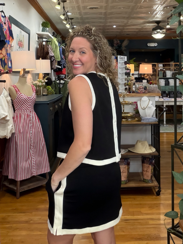 A woman wearing a black sleeveless dress with white contrast trim stands in a clothing boutique, looking back over her shoulder. The dress has a structured silhouette with a V-neckline and side pockets.