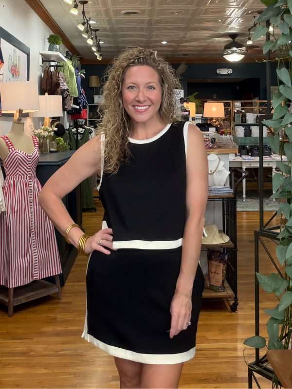 A woman with curly blonde hair stands in a boutique shop wearing a sleeveless black dress with white contrast trim at the neckline, waist, and hem. The background shows wooden floors, clothing racks, and store fixtures.