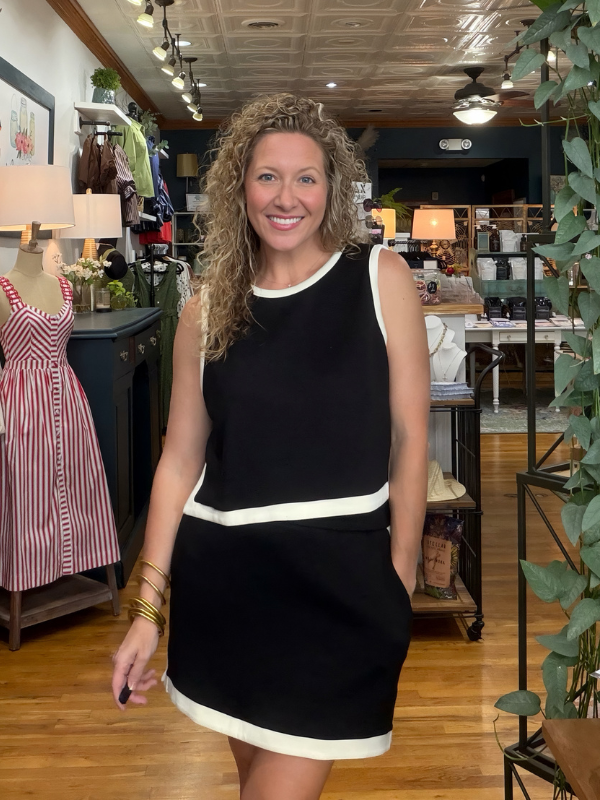 A smiling woman with curly hair stands in a boutique wearing a sleeveless black dress with contrasting white trim at the neckline, waist, and hem. The background shows a retail store interior with wooden floors, clothing racks, and plants.