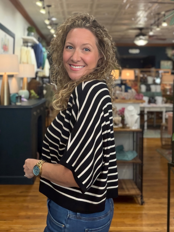 A smiling woman with curly blonde hair wearing a black and white striped knit top and blue jeans, standing in a retail store.