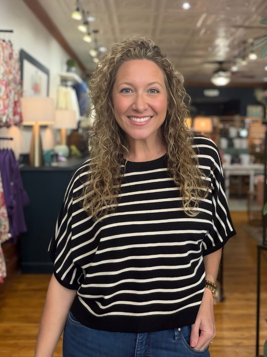 A smiling woman with curly blonde hair wearing a black and cream horizontal striped short-sleeve knit top and blue jeans, standing in a retail store.