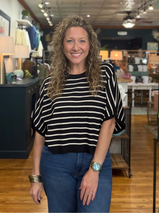 A smiling woman with curly blonde hair stands in a retail store wearing a black and white horizontally striped short-sleeve top and blue jeans.