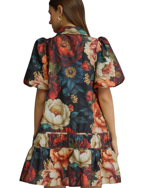 A woman shown from the back wearing a short-sleeve floral dress with puff sleeves and a tiered ruffled hem. The dress features a dark navy base with large red, cream, and blue floral prints.