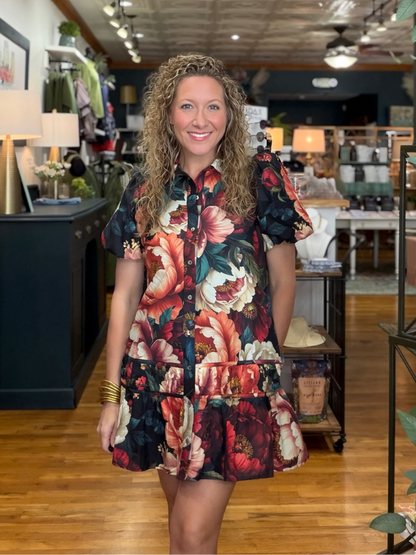 A woman with curly blonde hair stands in a boutique wearing a short-sleeve button-front dress with a bold floral print in red, pink, orange, cream, and dark green on a black background.