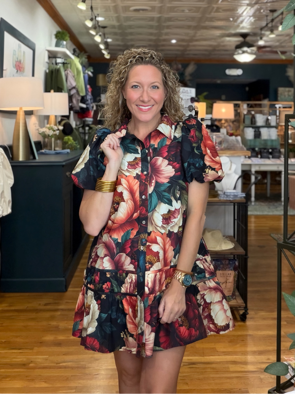 A woman with curly blonde hair stands in a boutique wearing a short-sleeve button-front dress with a bold floral print in red, pink, orange, and cream over a dark navy base. The dress features puffed short sleeves, a ruffled hem, and a tiered waist.