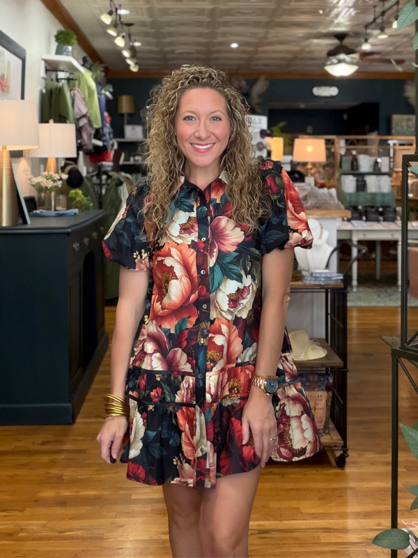 A woman with curly brown hair stands in a retail store wearing a short-sleeve button-up dress with a vibrant floral print in red, pink, and green on a dark background.