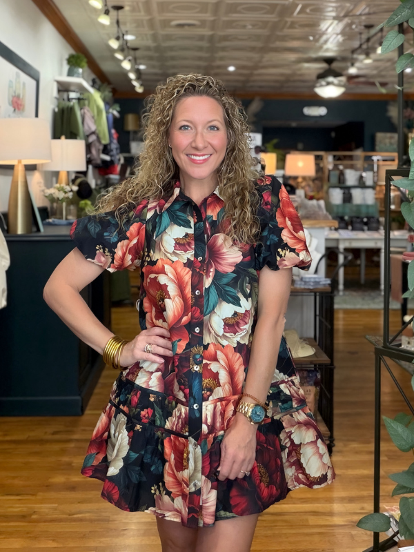 A woman with curly blonde hair stands in a retail store wearing a short-sleeve button-up dress with a vibrant red, orange, and teal floral print on a dark background.