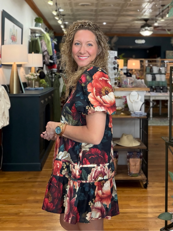 A woman with curly brown hair stands in a boutique shop, looking back over her shoulder. She is wearing a short-sleeve, knee-length dress with a dark navy background and a vibrant red, pink, and white floral print.