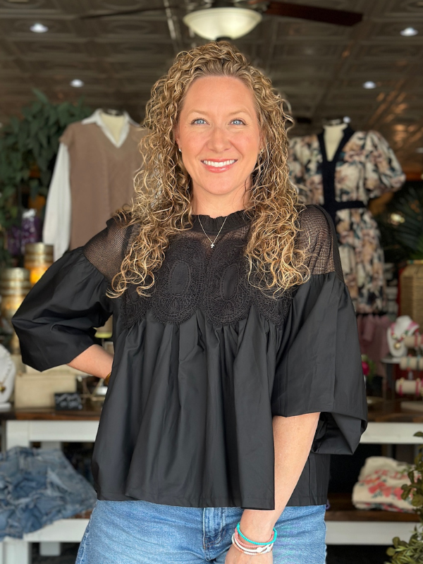 Woman wearing a black blouse with sheer sleeves and lace details in a store setting.