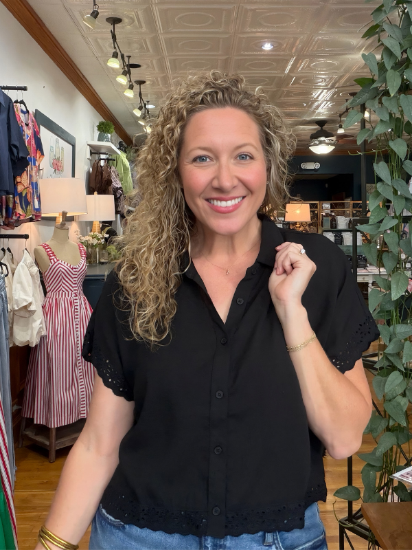 A smiling woman with curly blonde hair wearing a black short-sleeve button-up blouse with eyelet lace trim, standing inside a boutique store.
