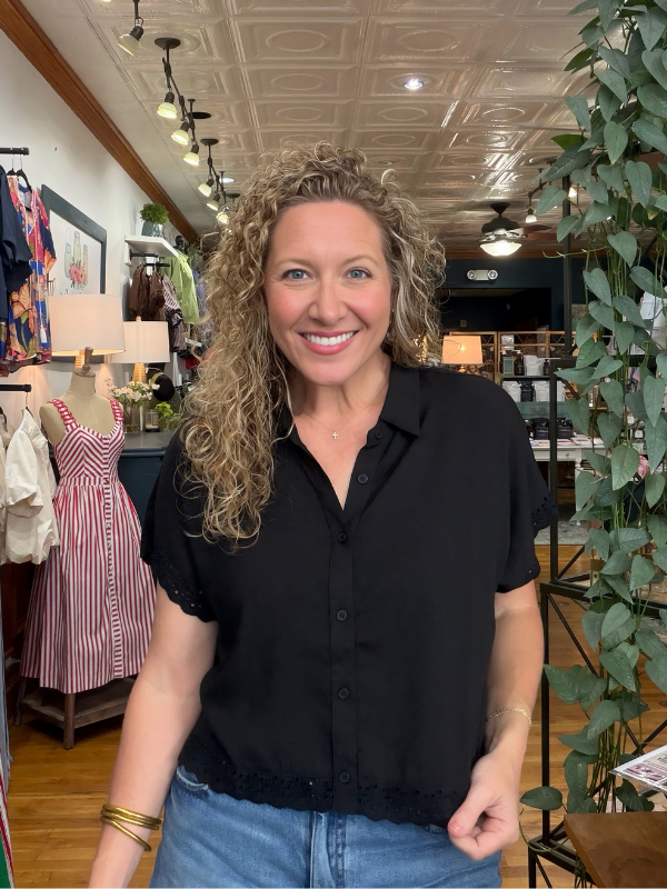 A smiling woman with curly blonde hair wearing a black short-sleeve button-up blouse and blue jeans, standing inside a retail boutique with clothing racks, plants, and store fixtures visible in the background.