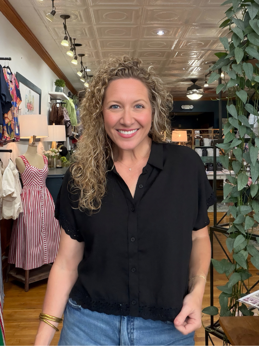 A smiling woman with curly blonde hair wearing a black short-sleeve button-up blouse and blue jeans, standing inside a retail boutique with clothing racks, plants, and store fixtures visible in the background.