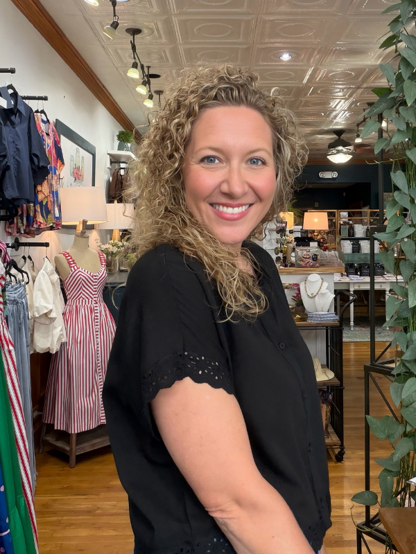 A smiling woman with curly blonde hair wearing a black short-sleeve blouse with eyelet lace trim, standing inside a retail boutique with clothing racks, lamps, and a decorative ceiling visible in the background.