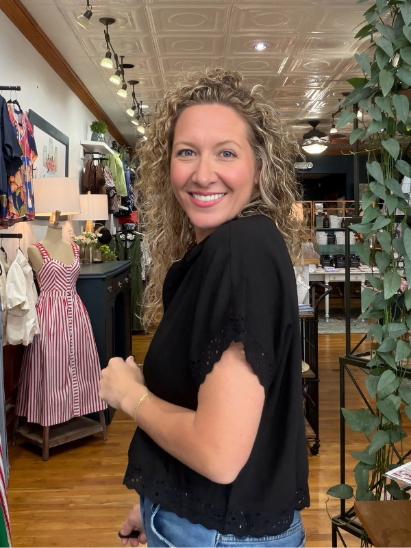 A smiling woman with curly blonde hair wearing a black short-sleeve eyelet top and blue jeans, standing inside a boutique store.