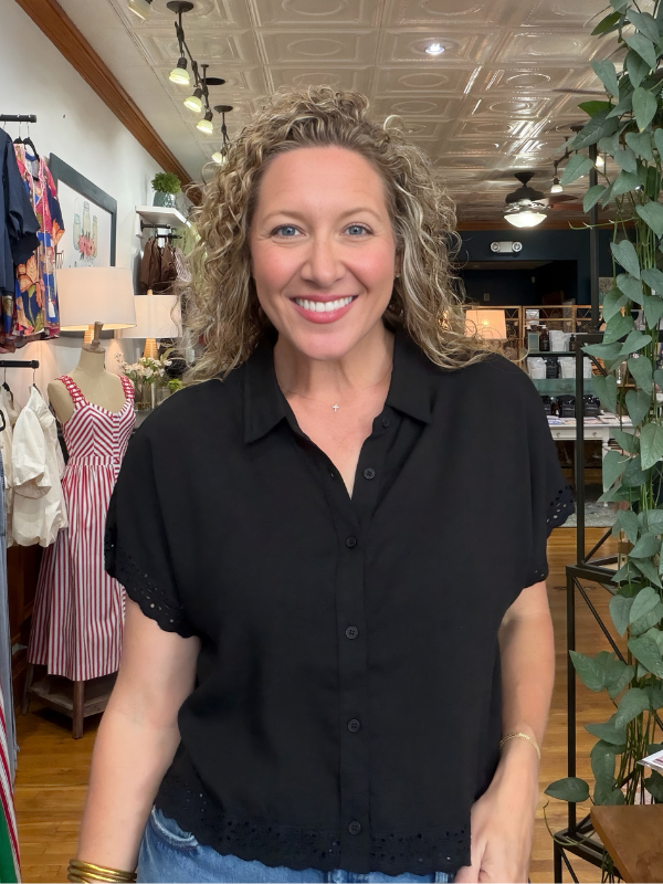 A smiling woman with curly blonde hair wearing a black short-sleeve button-up blouse with lace trim, standing inside a boutique store.