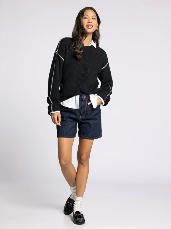 Woman wearing a black sweater with white accents, denim shorts, and white socks on a white background