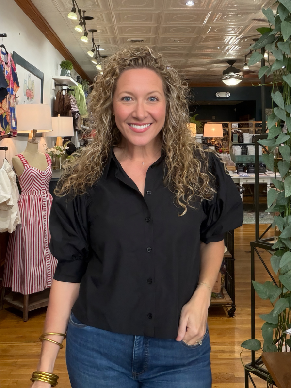 A smiling woman with curly blonde hair stands in a boutique wearing a black puff-sleeve button-up shirt and blue jeans.