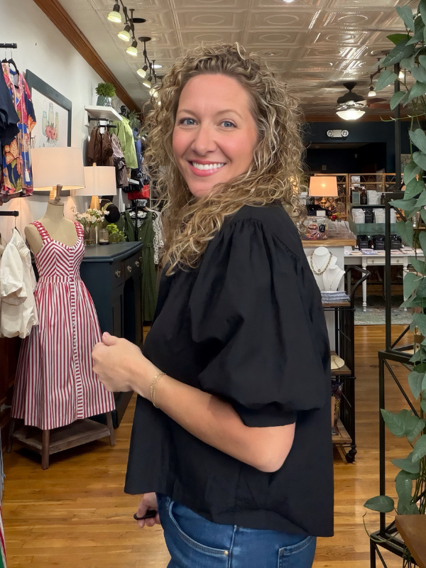 A smiling woman with curly blonde hair stands inside a boutique shop, wearing a black puff-sleeve blouse and blue jeans.