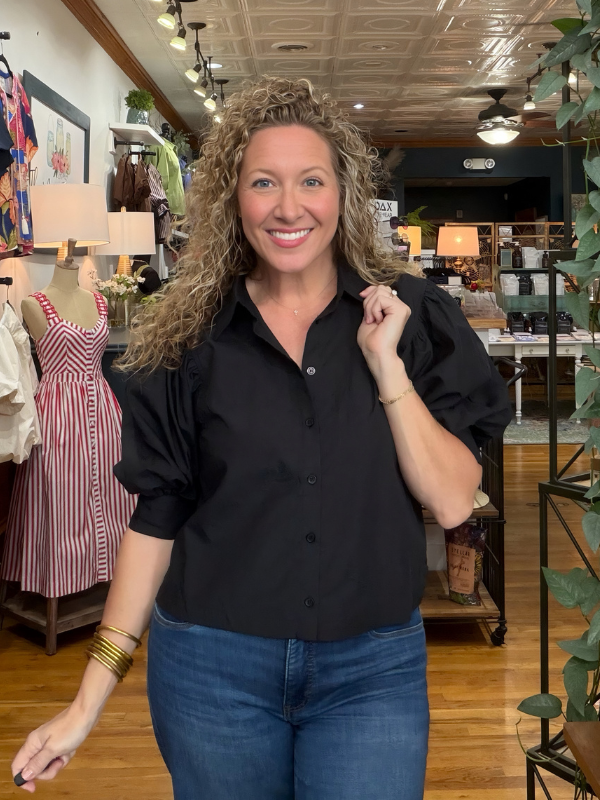 A woman with curly blonde hair wearing a black puff-sleeve button-down shirt and blue jeans, standing inside a retail boutique.