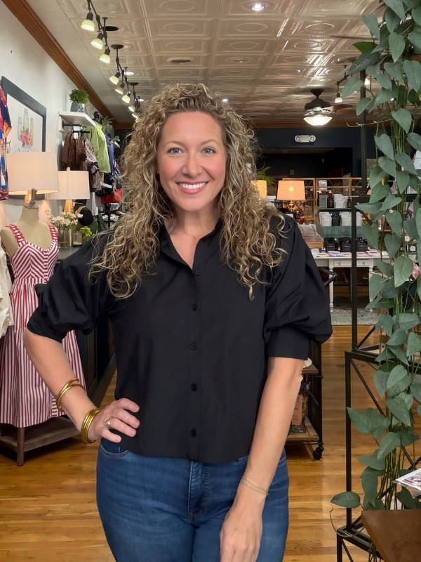 A smiling woman with curly hair stands in a boutique wearing a black short-sleeve button-up blouse with puff sleeves and blue jeans.