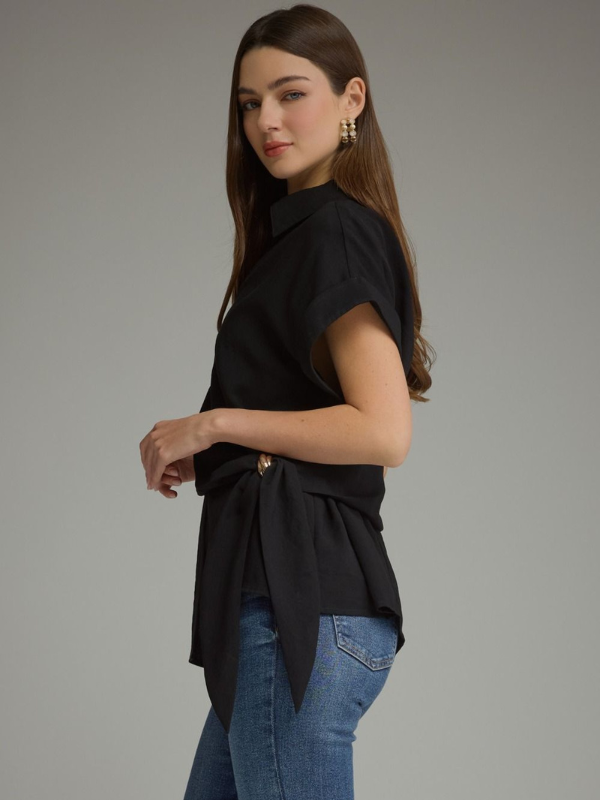 A woman modeling a black short-sleeve collared blouse with a side tie detail, paired with blue jeans, against a solid gray studio background.