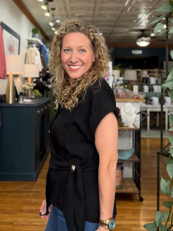 A smiling woman with curly blonde hair stands in a boutique shop, wearing a black short-sleeve wrap-style top with a tie detail at the waist and blue jeans.