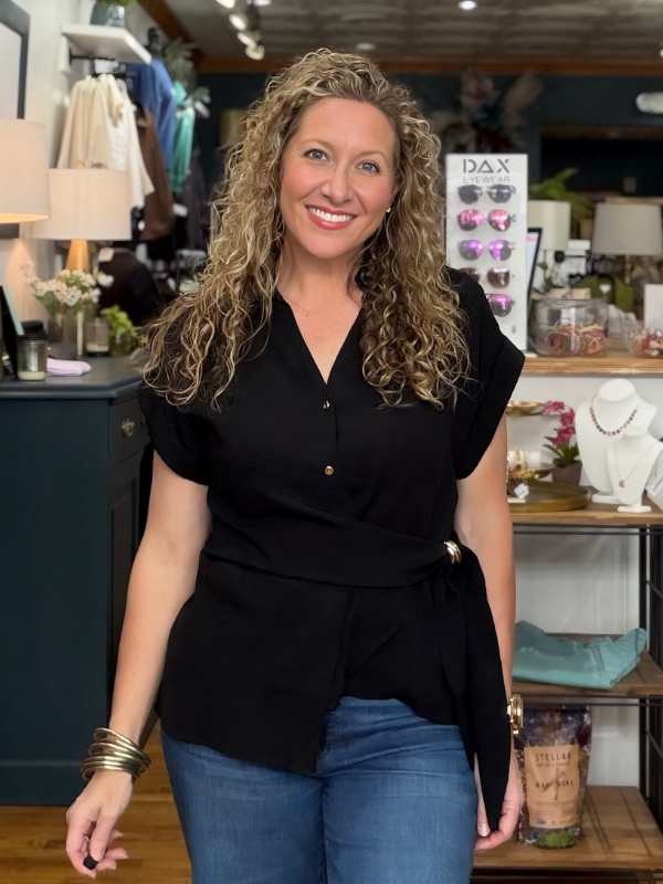 A smiling woman with curly blonde hair wearing a black short-sleeve button-front top and blue jeans, standing inside a retail store.