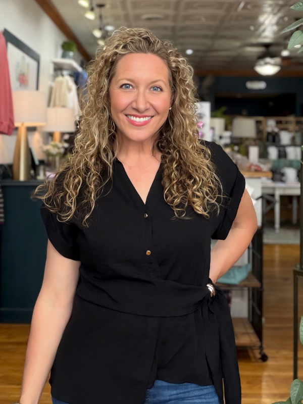 A smiling woman with long curly blonde hair stands in a retail store wearing a black short-sleeve button-front top with a side tie detail and blue jeans.