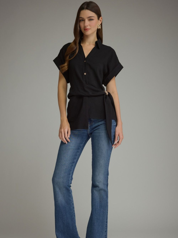 A woman modeling a black short-sleeve button-front blouse with a self-tie waist sash, paired with blue jeans, against a solid gray studio background.