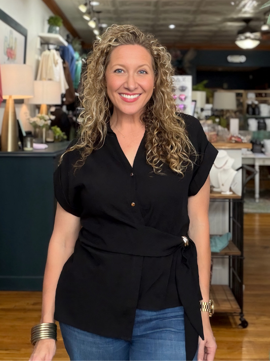 A smiling woman with curly blonde hair wearing a black short-sleeve wrap-style blouse with a tie waist and blue jeans, standing inside a retail store.
