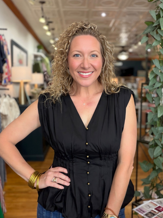 A smiling woman with curly blonde hair wearing a sleeveless black button-front top, standing in a boutique interior with clothing racks and plants in the background.
