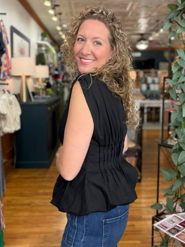 A woman with curly blonde hair wearing a sleeveless black peplum top and blue jeans, posing inside a retail store.