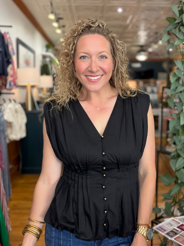 A smiling woman with curly blonde hair wearing a sleeveless black button-front top and blue jeans, standing inside a retail store.