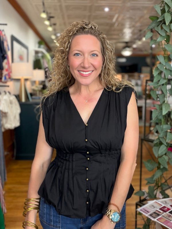 A smiling woman with curly blonde hair wearing a sleeveless black button-front top and blue jeans, standing inside a boutique store with clothing racks, lamps, and greenery in the background.