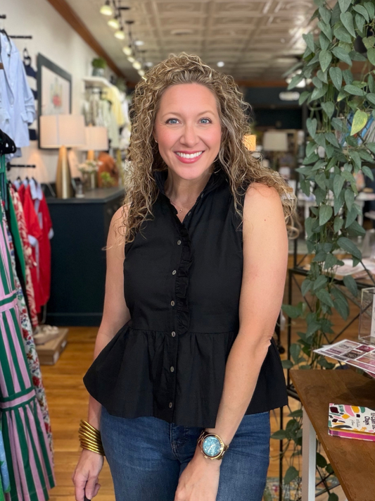 Woman in a black sleeveless top and blue jeans standing in a store with plants and clothing racks.