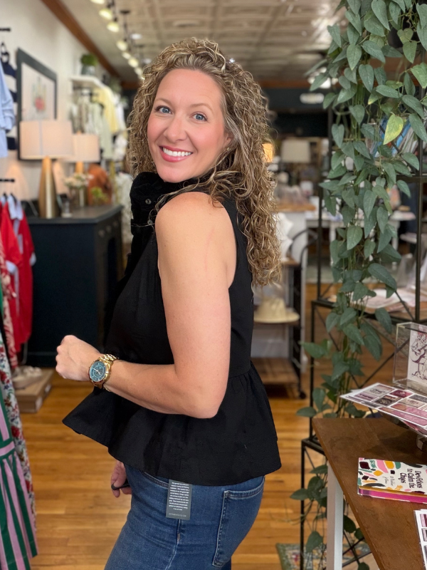 Woman in a black sleeveless top and blue jeans standing in a store with plants and furniture.