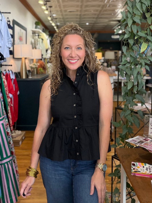 Woman in a black peplum top and blue jeans standing in a store with plants and clothing in the background.