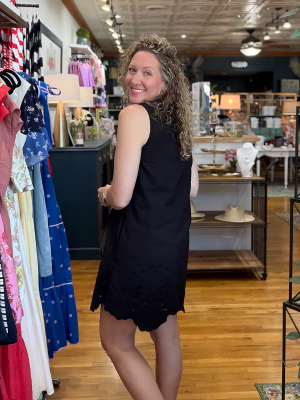 Woman in a black dress standing in a clothing store with racks of clothes and a counter in the background.