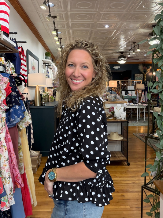 Woman in a polka dot blouse standing in a store with clothing racks and decor.
